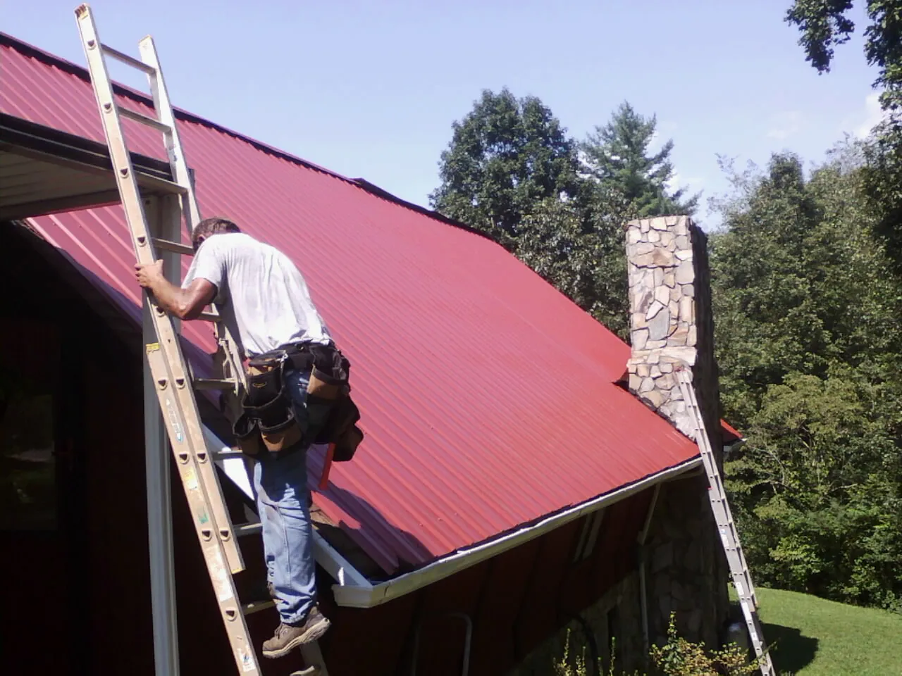 Roofer on a ladder beside a red metal roof and stone chimney — Soffit & Fascia Repair by Roofing Brothers in Blomkest